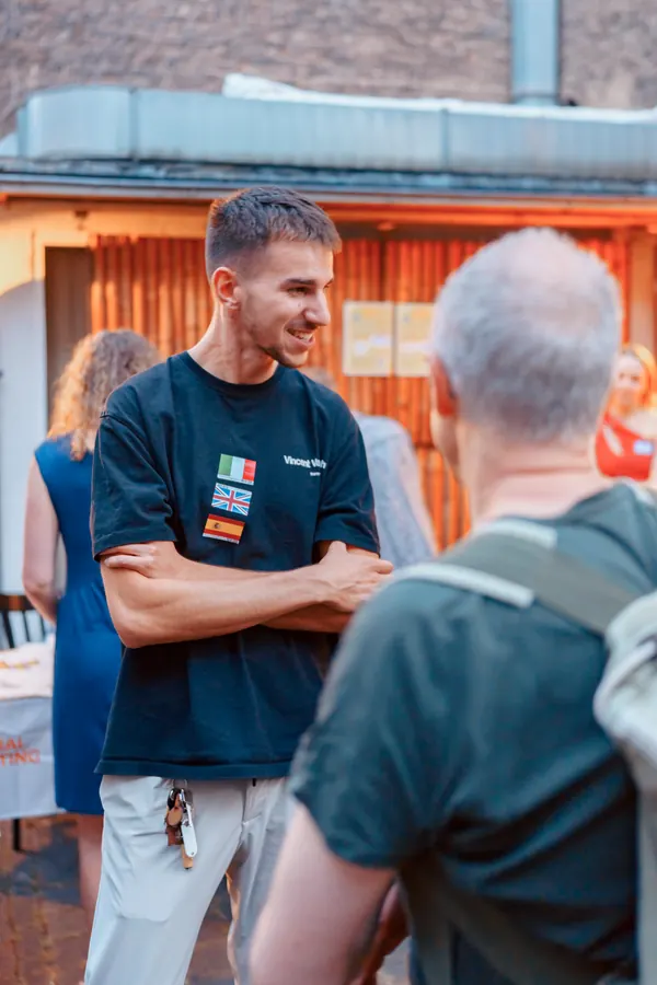 A guest with flag stickers smiling in conversation in the courtyard at the SMP Thursday social in Bonn