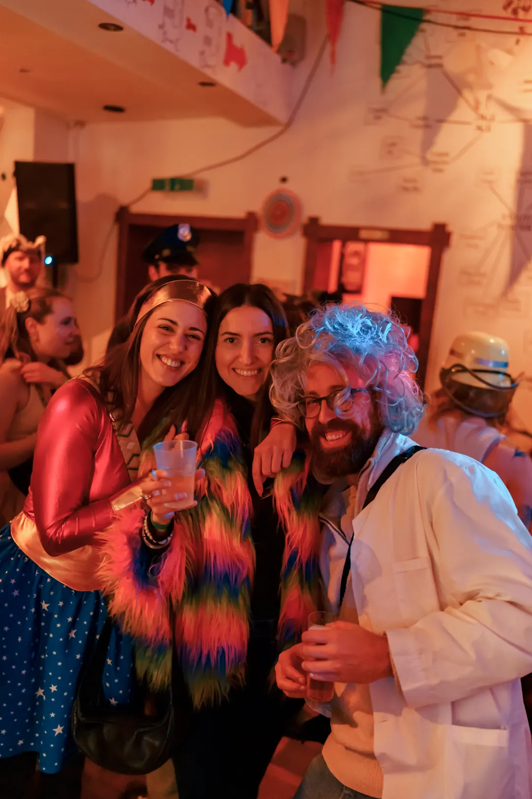 Two friends in carnival costumes and a man in a white wig and lab coat smiling together at Craftbeer Corner Coeln on 11.11
