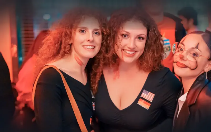 Three friends in Halloween face paint smiling with flag stickers at a Social Melting Pot night in Cologne