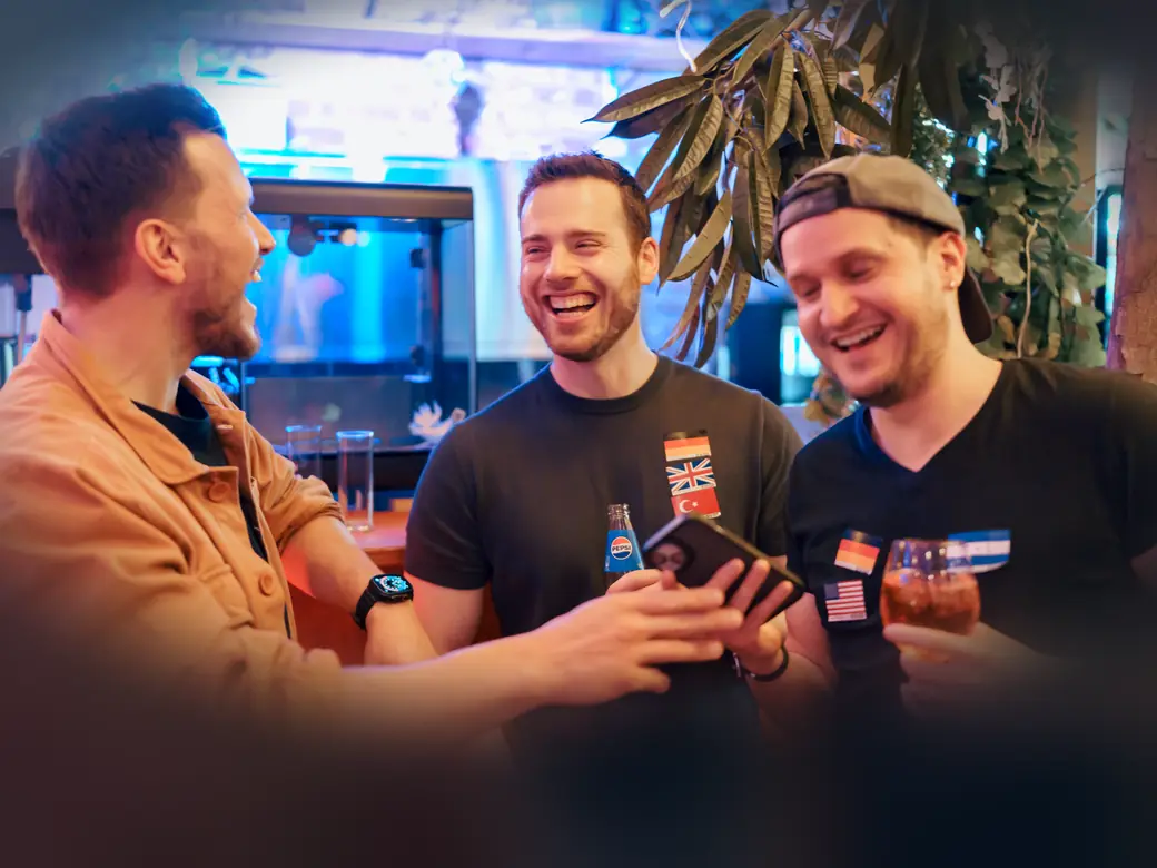 Three guys laughing at the bar while one shows the others his phone at a Social Melting Pot night in Cologne
