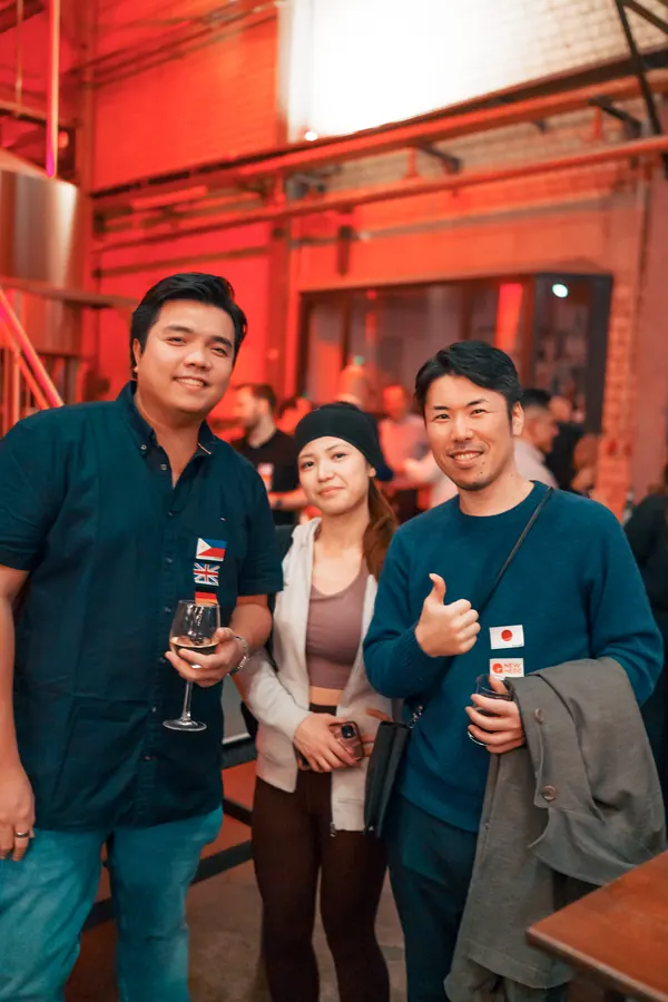 Three international guests with flag stickers smiling in the brewery hall at the SMP social in Düsseldorf