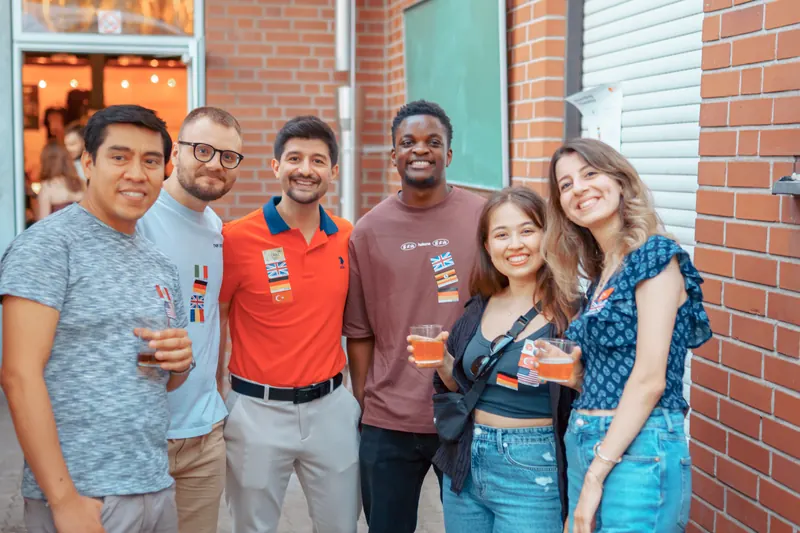 Six guests with language flag stickers smiling outside Brauerei Kürzer at the SMP social in Düsseldorf