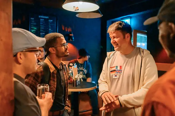 Three guests with language flag stickers laughing at a table inside Bar Grüner Jäger at the SMP social in Hamburg