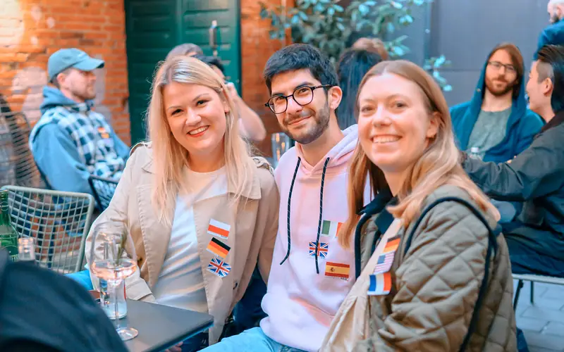 Three guests with language flag stickers smiling at a terrace table at the Wednesday social in Hamburg