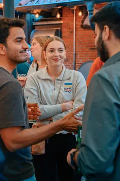 A guest with language flag stickers smiling as two others start a conversation at the Wednesday social in Hamburg