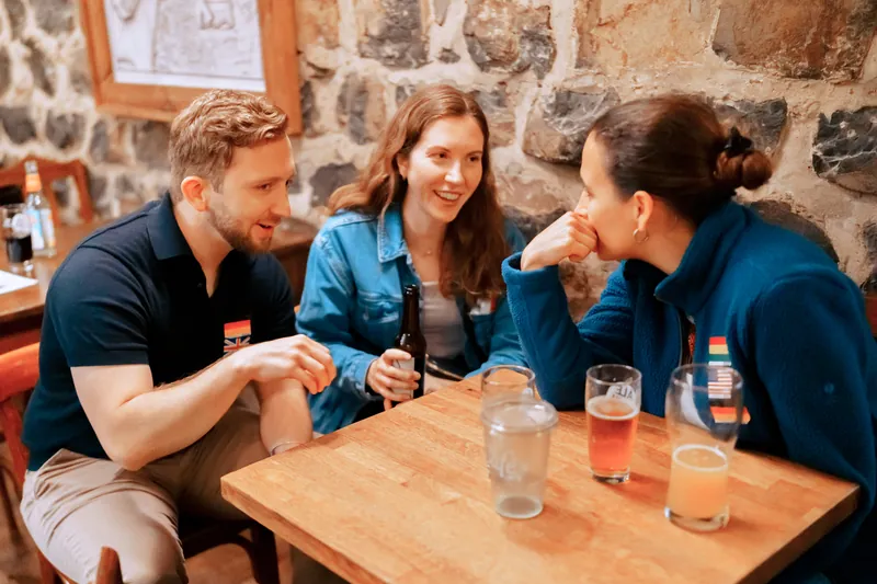 Three guests with language flag stickers talking and laughing at a table inside °Plato Beer & BBQ in Bonn